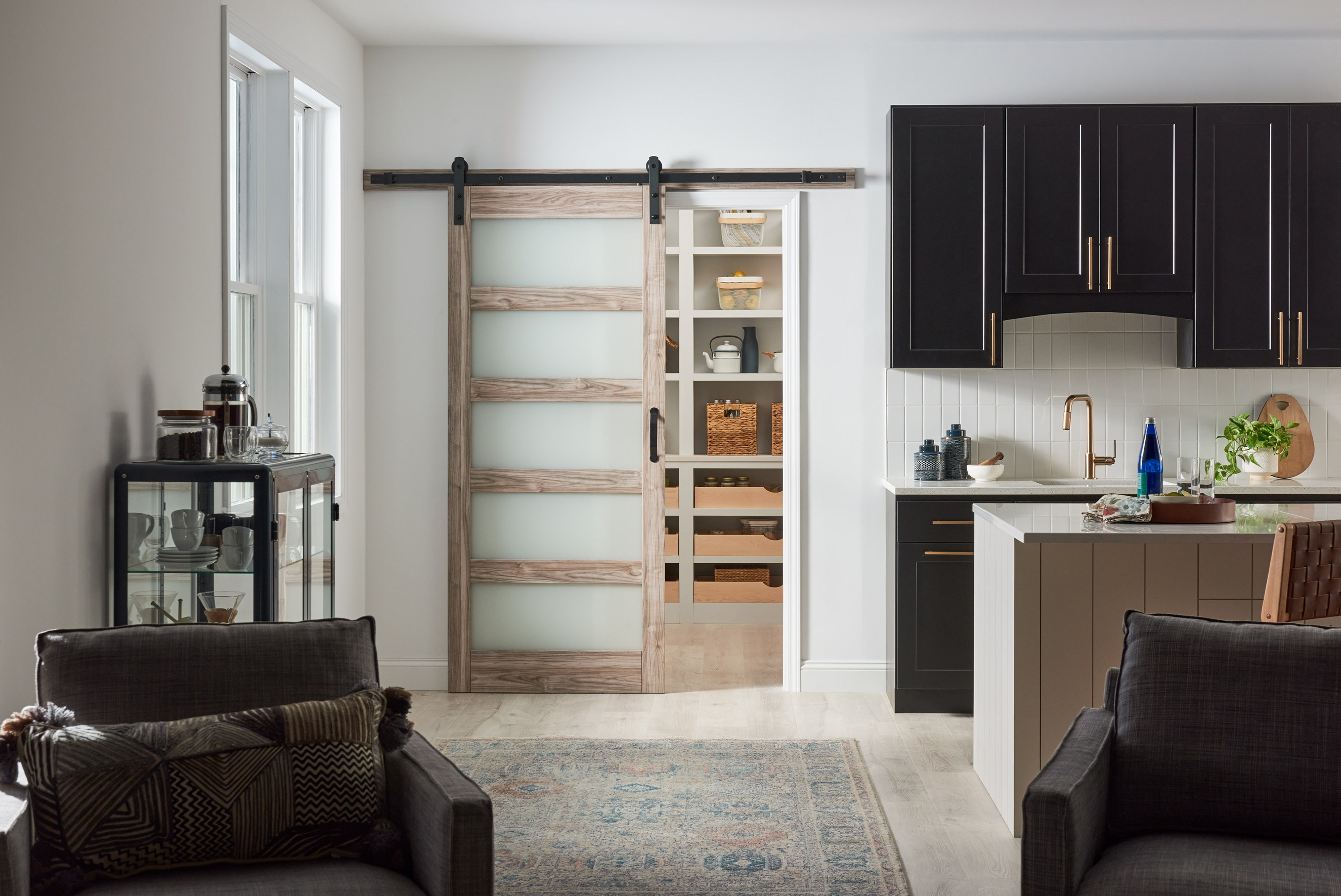 Front view of a transitional style lite ash grey 5-lite barn door with glass inserts leading to a pantry in a kitchen with black cabinets. There is a carpet on the wooden floor behind two dark brown cloth armchairs.